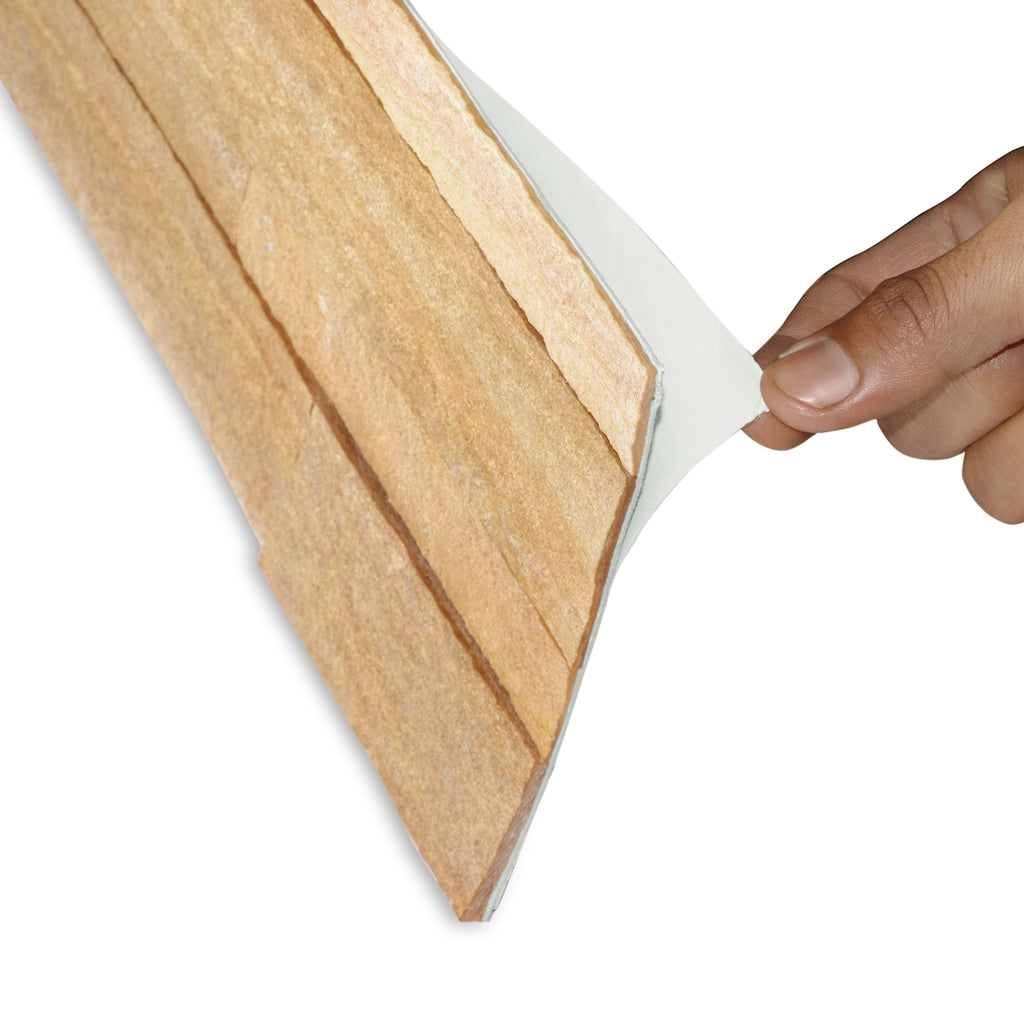 A product image of light brown colored burly wood peel and stick self-adhesive real stone wall tiles from stoneflex shown sideways with a hand peeling the protective film off its self-adhesive back.